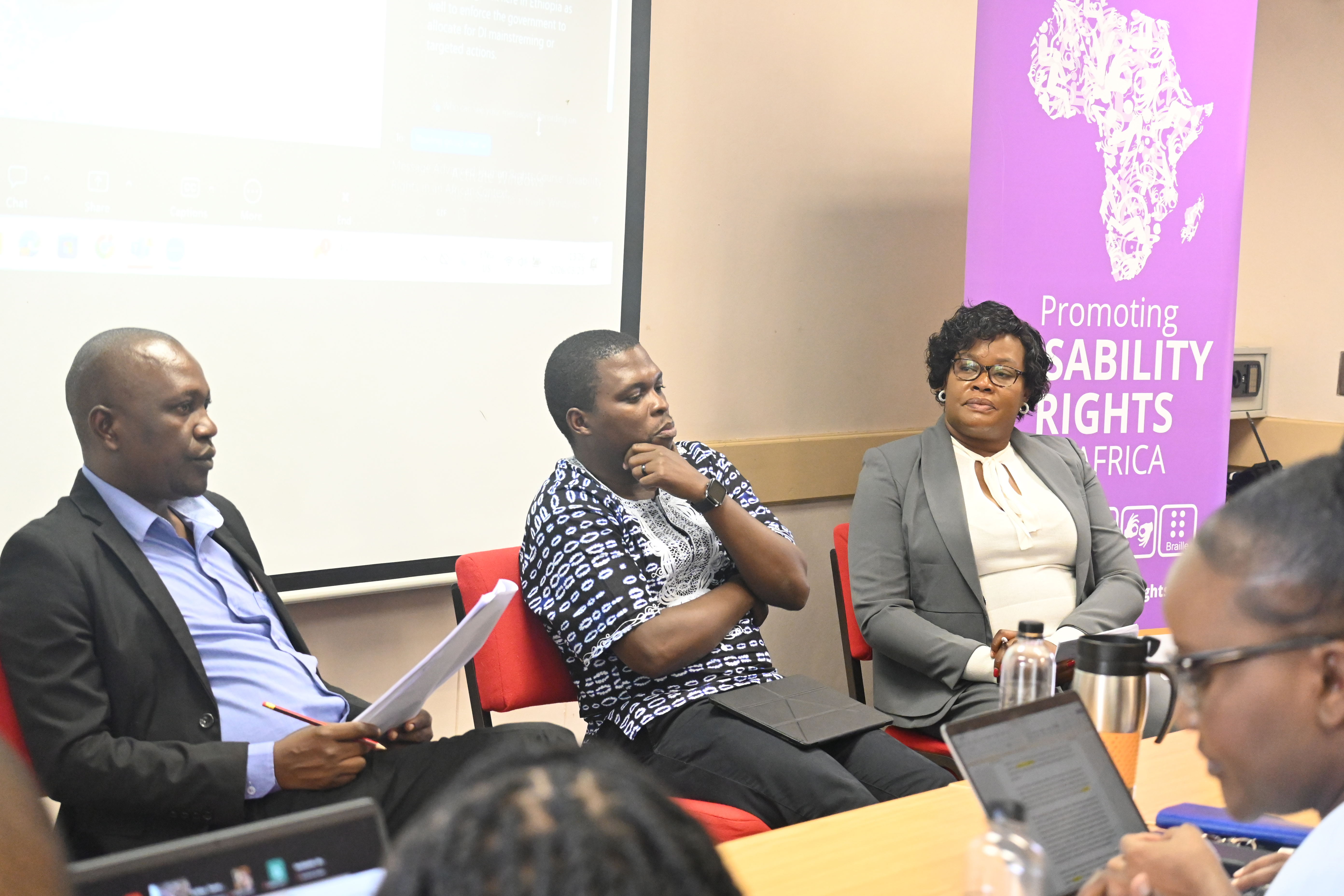 Image 1 - Moderator and panelists (Esau Mandipa, Phakama Shili and Shishemwbe Mwale) seated at the front of the classroom. In the image, the panelists appear to be listening to and reflecting on engagements from the audience. The Disability Rights Unit banner is also visible in the image. The banner is purple with white writing.