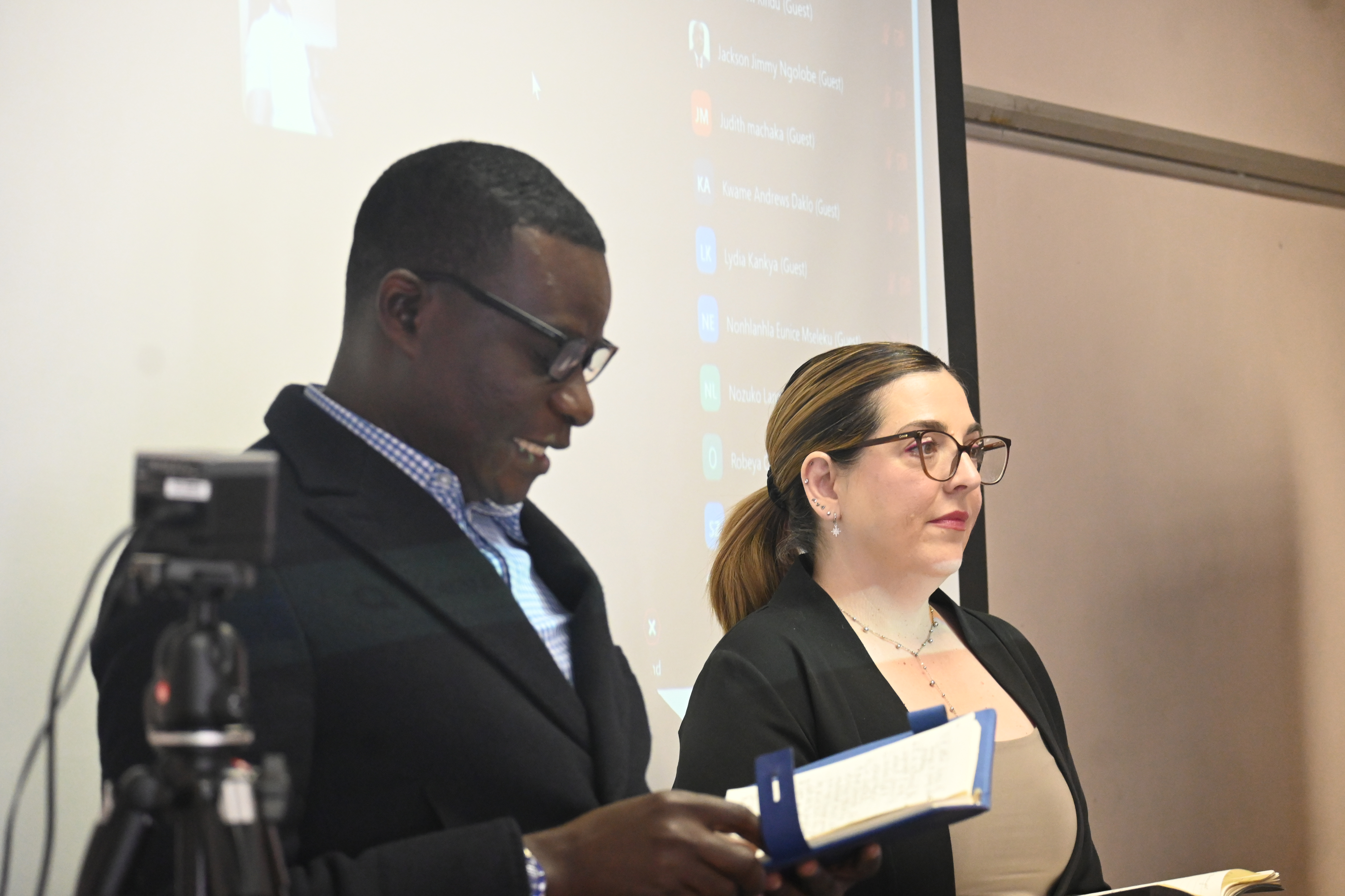 Image 3 - Phillip Sabuni and Jahni De Villiers standing at the front of the classroom delivering the recap from the previous days' sessions. They are both dressed in black blazers. The screen appears behind them. In the image, there is also a tripod next to Phillip.